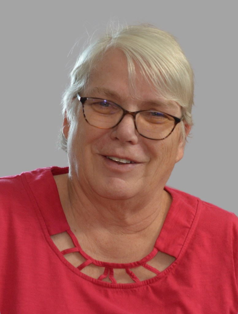 Image of Connie smiling. Connie has white hair in a pixie cut. She is wearing dark cat eye glasses and a fuchsia top with cut outs around the neck.