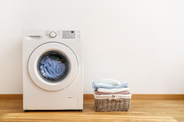 A laundry room with white walls and light wooden floor. There is a front load washing machine withe blue cloth in it. There is a wicker basket on the side with nicely folded clothes in blues and neutrals colours.